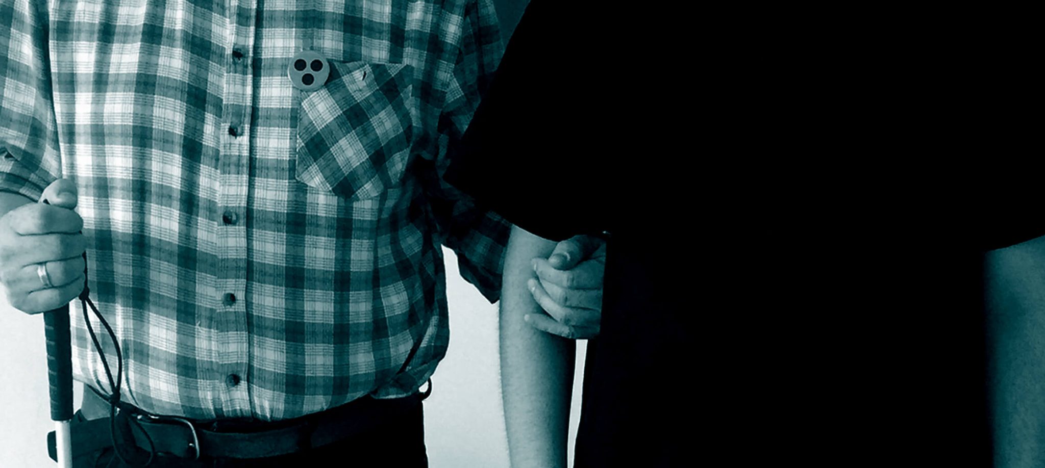 Eine Person mit Blindenstock und einem Button mit dem Blinden-Symbol am Hemd steht eingehängt mit einer anderen Person.