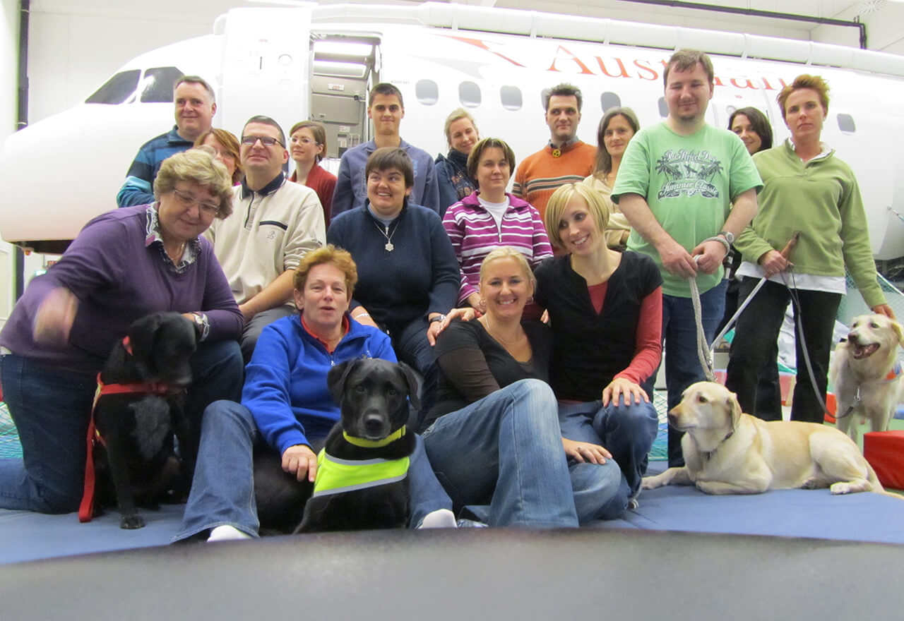 Eine Gruppe von Menschen – zum Teil mit Assistenzhunden – vor einem Flugzeug der Austrian Airlines.