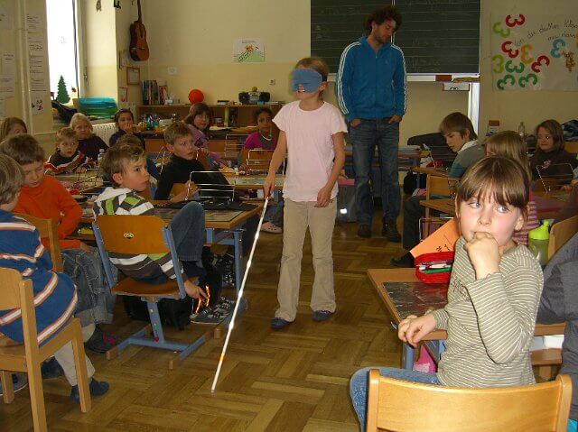 Ein Kind mit Dunkelbrille und Blindenstock geht durch die Klasse.