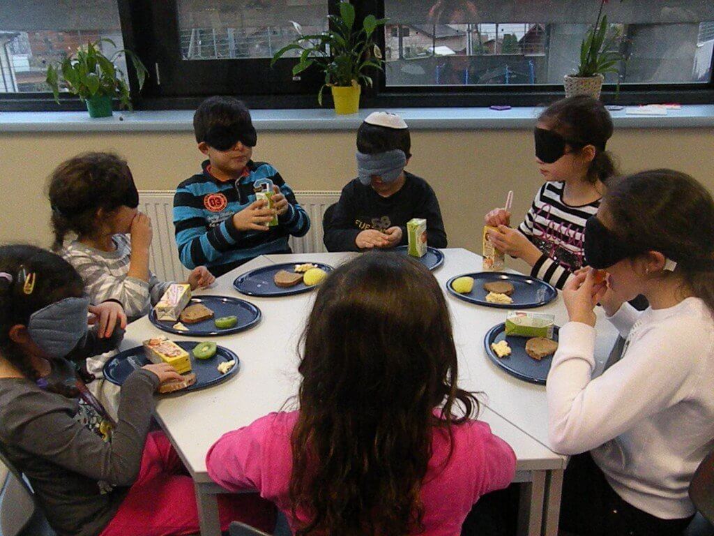 Kinder mit Dunkelbrille sitzen um einen sechseckigen Tisch und essen Jause