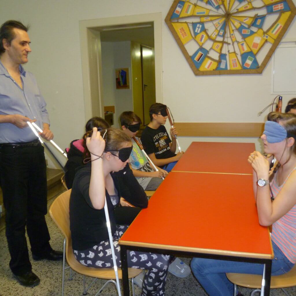 SchülerInnen mit Blindenstock und Dunkelbrille sitzen um Tisch während Harald etwas erklärt