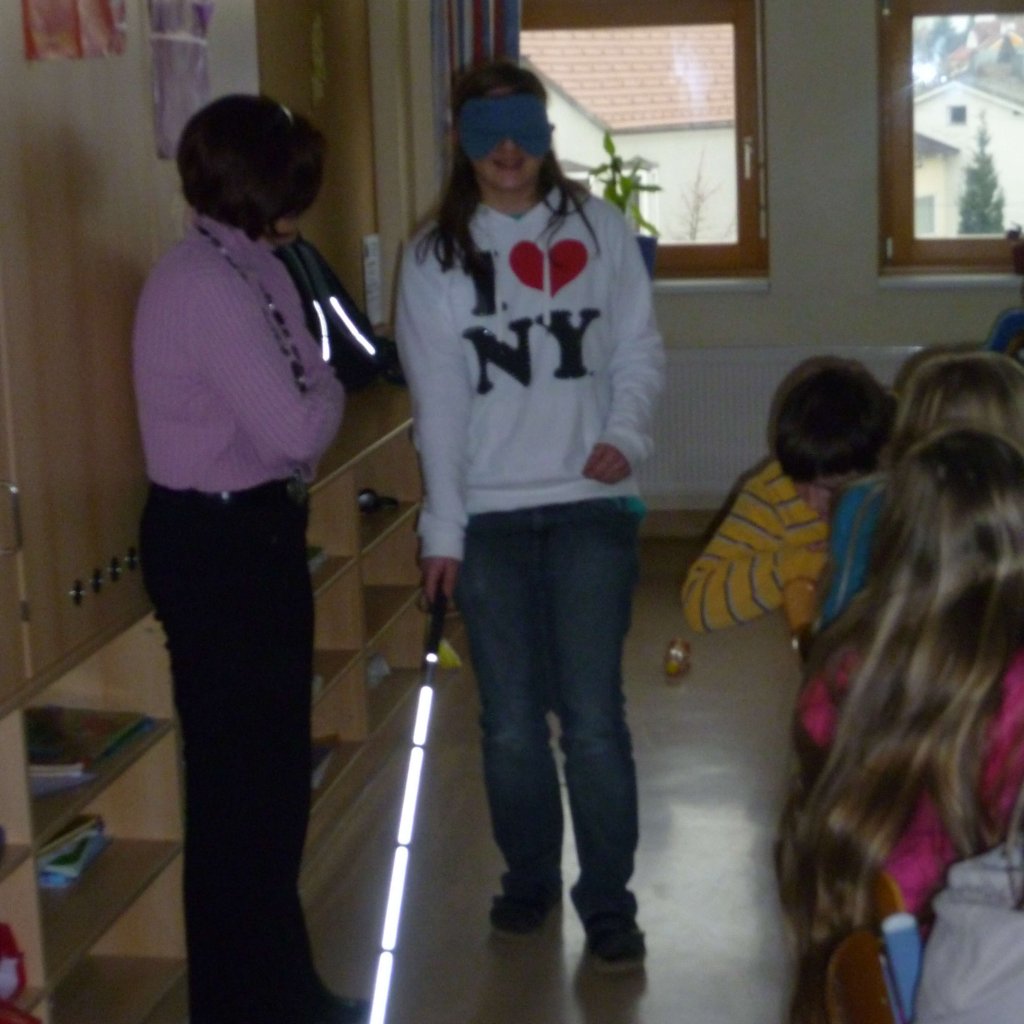 Schülerin geht mit Blindenstock und Dunkelbrille zwischen Lehrerin und Sitzreihe