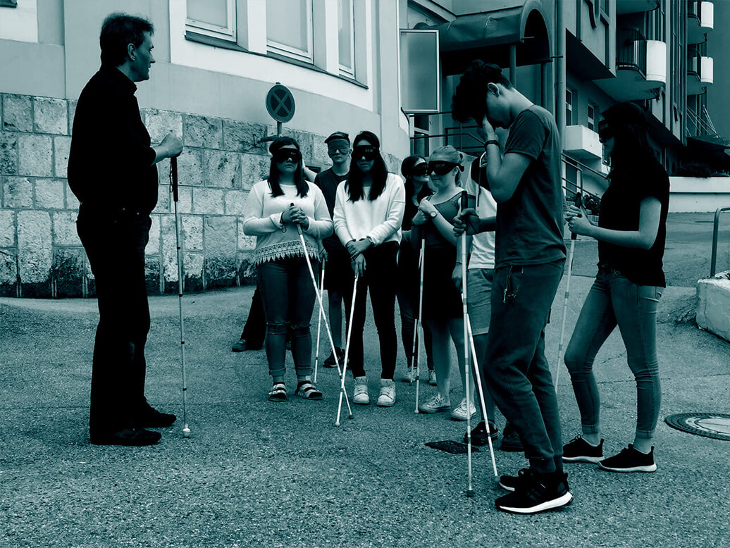 Harald steht vor einer Gruppe SchülerInnen, die SchülerInnen haben Dunkelbrillen auf und einen Blindenstock in der Hand.