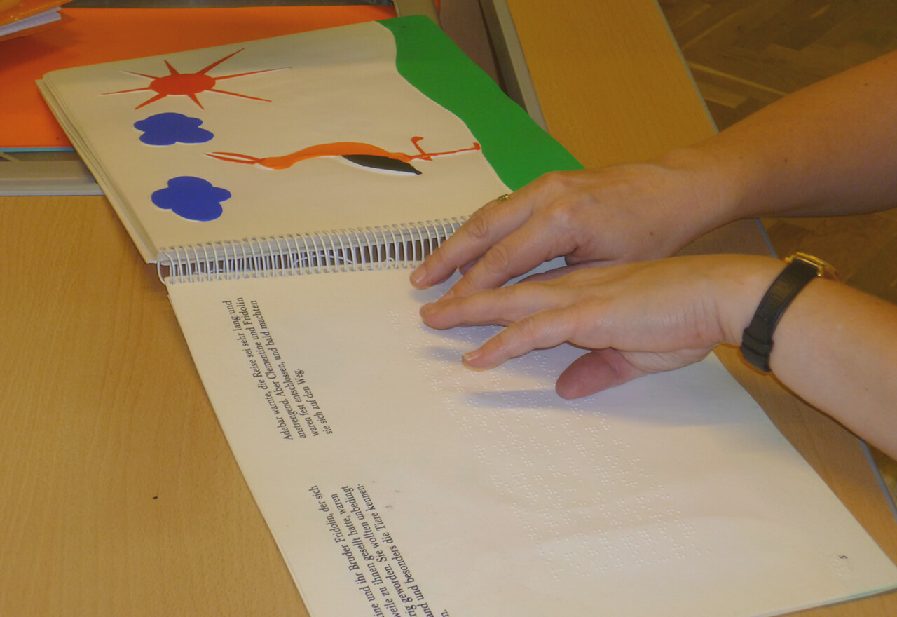 Sabine liest Braille Kinderbuch, man sieht ihre Hände auf dem Buch