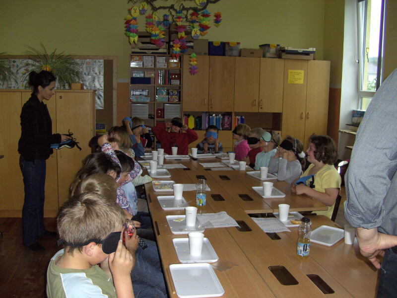 Alle Kinder sitzen an einem großen Tisch. Vor ihnen ein Tablett mit Restbrösel eines Essens und einem Trinkbecher.
